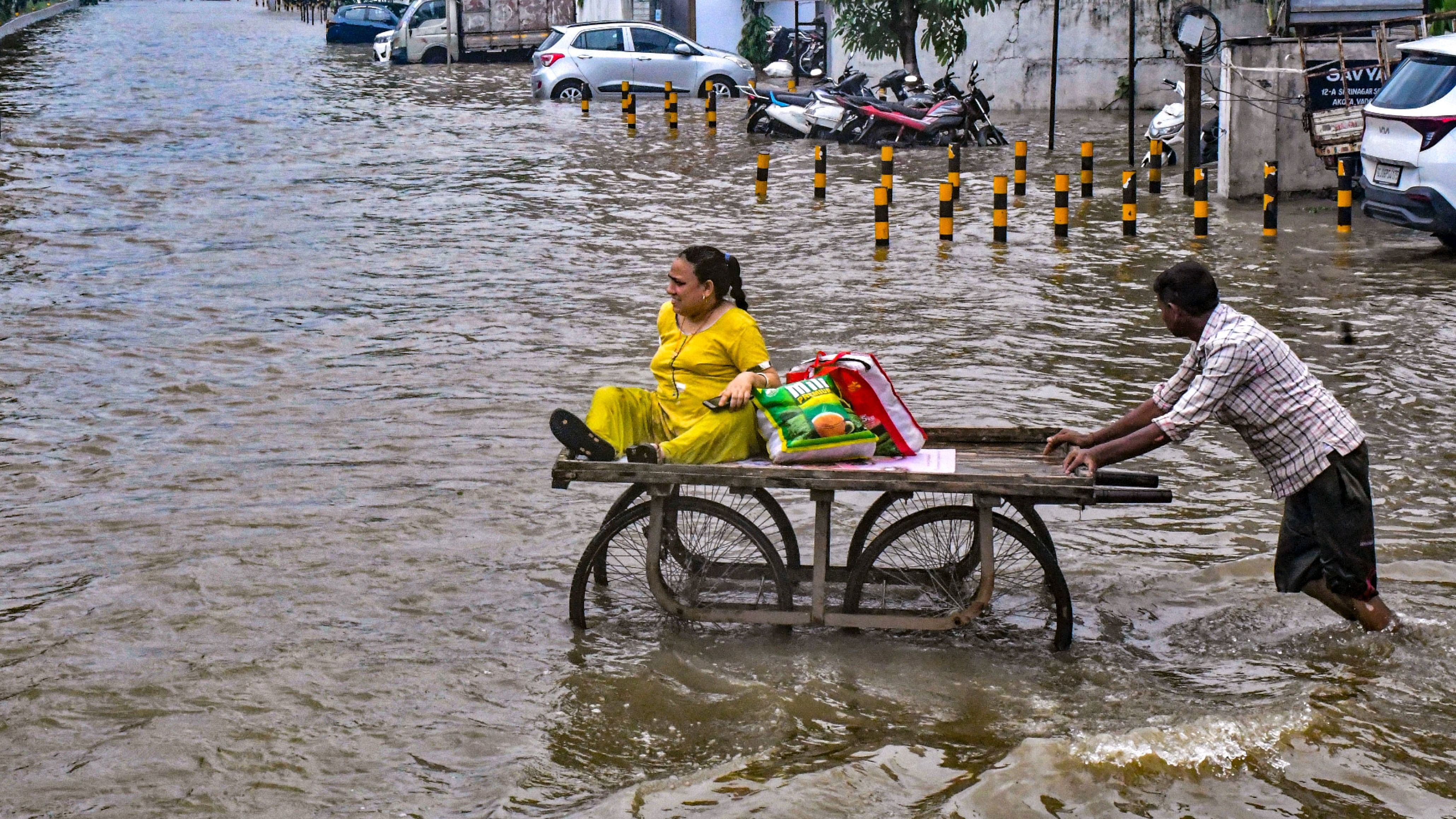 <div class="paragraphs"><p>A labourer pushes a cart carrying a woman to help her cross a flooded road after heavy monsoon rainfall, in Vadodara, Tuesday.</p></div>